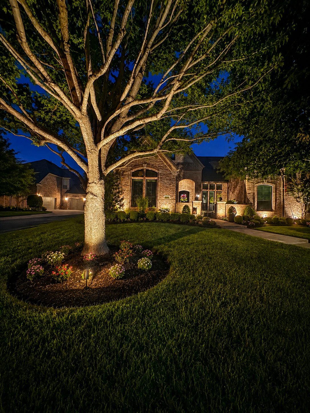 Brick home at night with tree uplighting in a flower bed and architectural lighting on the brick
