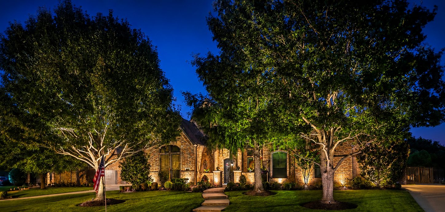 Brick home from the street with a flag, tree uplighting on two trees, and architectural lighting on the brick