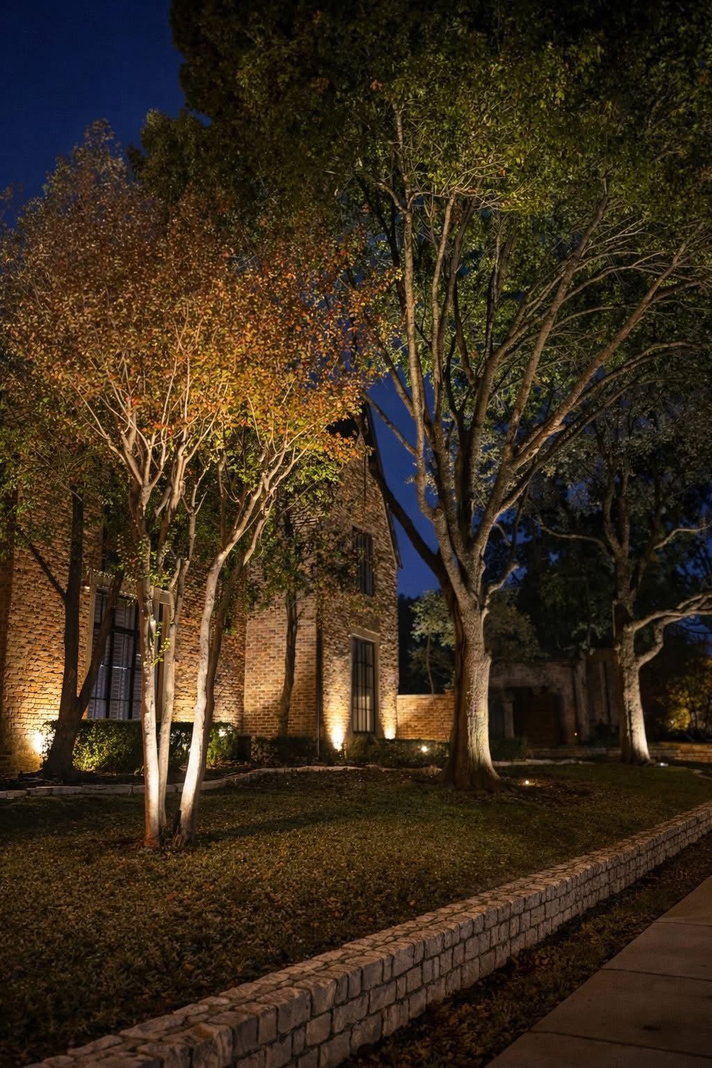 Architectural lighting on brick, with tree uplighting beside a low stone wall