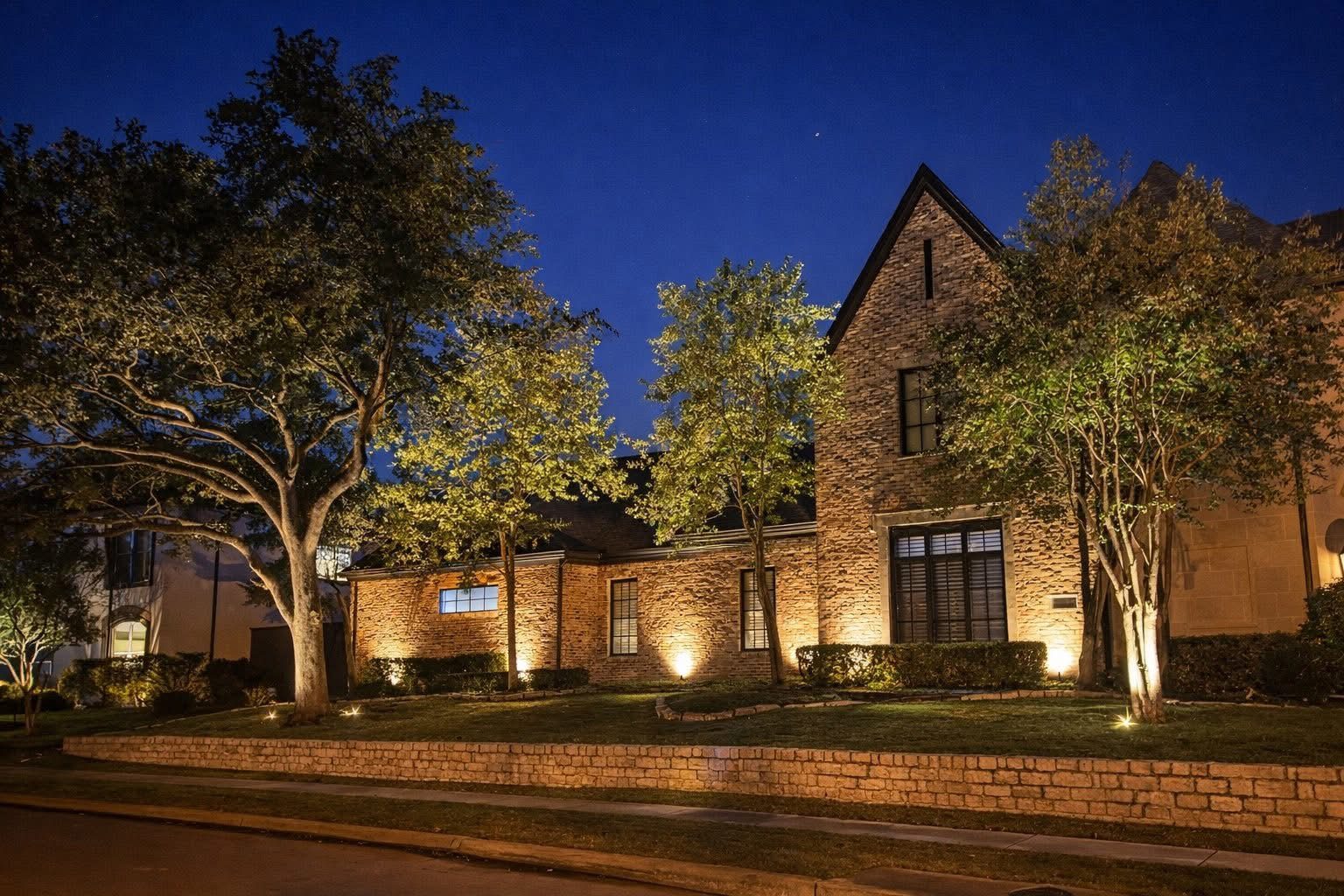 Architectural lighting on a brick home, with tree uplighting along a low stone wall