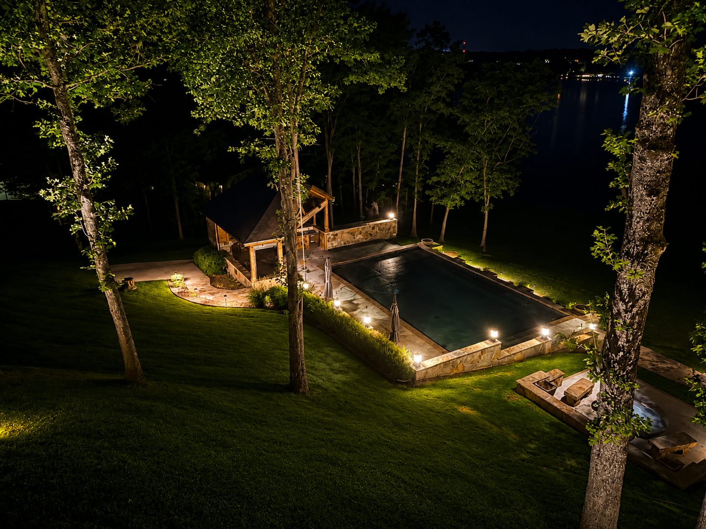 Aerial view of a pool, pavilion, and lake, with tree uplighting
