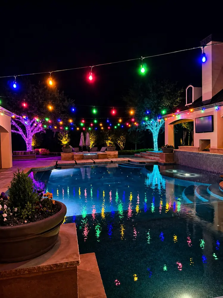 Pool at night with multicolor string lights reflected in the water, wrapped trees, and an outdoor kitchen