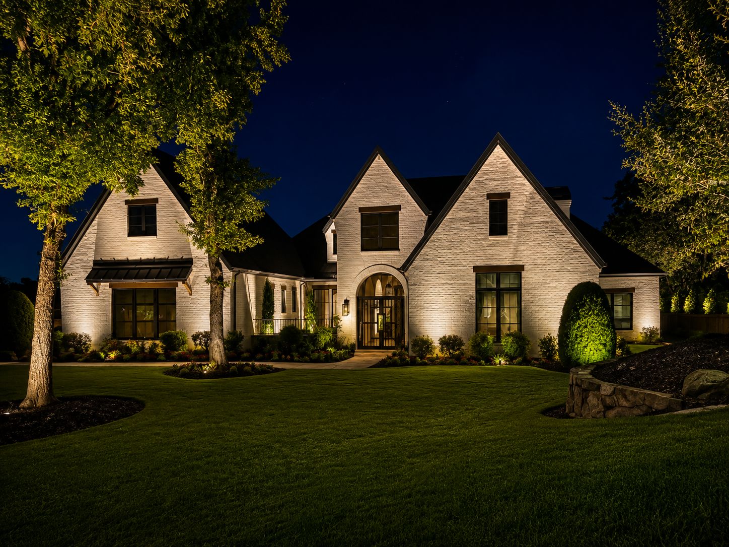 White-brick home at night with gables, warm uplighting on the brick, and tree uplighting