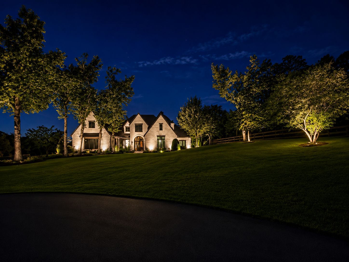Wide night view toward a white-brick home and tree uplighting