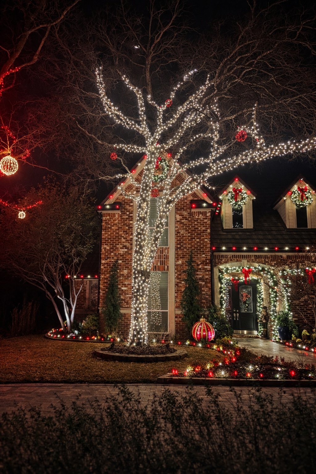 Wrapped tree with Christmas ornaments in front of a brick home with wreaths and garland