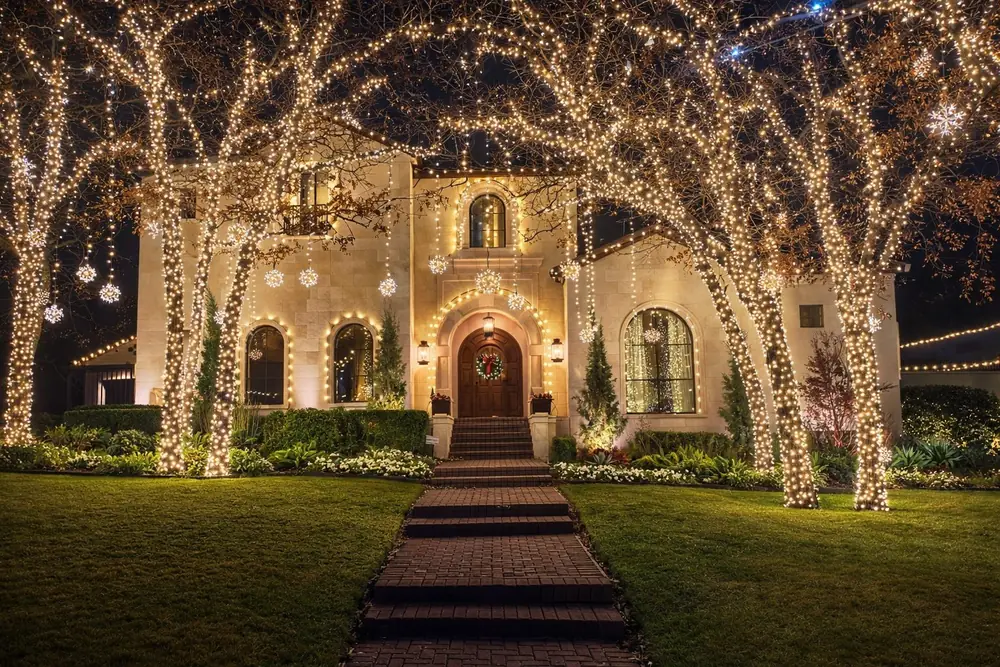 Christmas lighting with wrapped trees and a brick walk to an arched door with a wreath